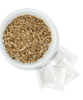 A bowl filled with dried burdock root next to several tea bags on a white background.