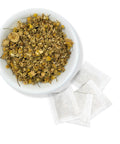 A white bowl filled with dried chamomile flowers, alongside individual chamomile tea bags.