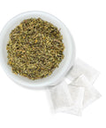 White bowl filled with dried herbs next to white tea bags on a white background