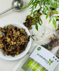 Evergreen Herbs Flor de Manita dried flowers in a white bowl with a tea infuser and product pouch beside it.