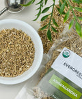 Evergreen Herbs Ashwagandha dried cut roots displayed in a white bowl and a branded pouch, with a tea infuser and green leaves on a clean white surface.