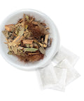 A bowl of dried herbal tea ingredients including pieces of bark, leaves, and roots with tea bags beside it.