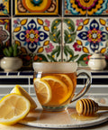 Glass mug of hot tea with lemon slices, honey dipper, and a cut lemon on a tiled countertop in front of colorful decorative tiles.