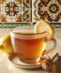 A glass cup of tea garnished with a lemon slice, with fresh lemons and a honey dipper placed nearby on a kitchen counter with colorful decorative tiles in the background.