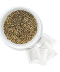 A white bowl filled with dried spearmint tea leaves, alongside two individual spearmint tea bags.